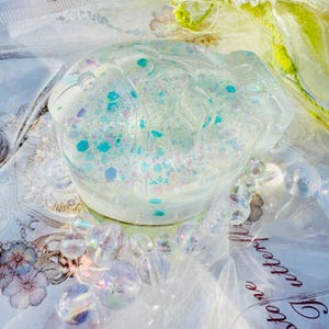 May include: A clear, round, resin paperweight with a glittery, iridescent finish. The paperweight is filled with blue, green, and pink glitter.