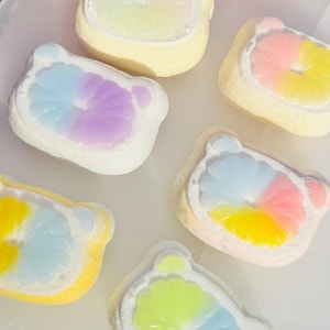 May include: An overhead shot of several colorful, bear-shaped bath bombs. Each bath bomb has a white border and a colorful center, with shades of yellow, pink, blue, and purple. The bath bombs are arranged on a white surface.