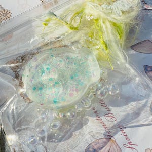 May include: A clear, iridescent resin charm shaped like a fish with a glittery, pastel blue, pink, and green design. The charm is surrounded by clear, iridescent beads and is sitting on a white surface with the text "Every stone... Butterfly" visible.