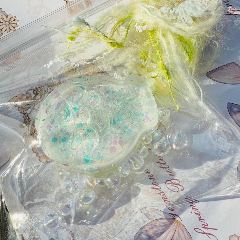 May include: A clear, iridescent resin charm shaped like a fish with a glittery, pastel blue, pink, and green design. The charm is surrounded by clear, iridescent beads and is sitting on a white surface with the text "Every stone... Butterfly" visible.