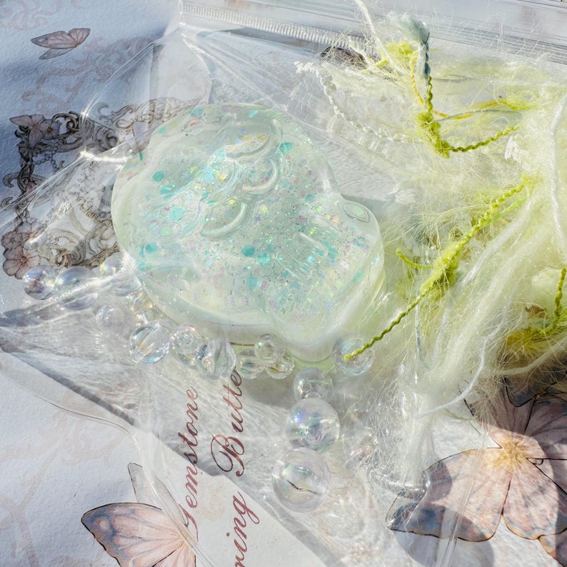 May include: A clear, iridescent resin skull filled with glitter and iridescent flakes. The skull is sitting on a white surface with a butterfly design.