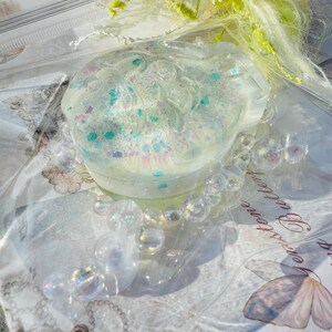 May include: A clear, round resin trinket box with a swirling, glittery design. The box is filled with iridescent, clear beads and is sitting on a white surface.