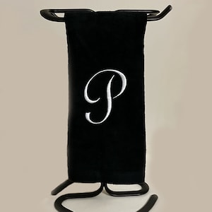 May include: A black hand towel with a white embroidered letter "P" is displayed on a black metal towel rack. The towel is folded and hanging vertically. The rack has a curved base and a bar to hold the towel.