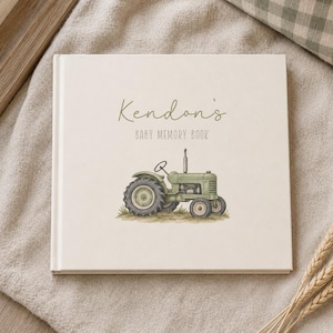May include: A square, off-white baby memory book featuring the text 'Kendon's BABY MEMORY BOOK' in dark green lettering. Below the text is a detailed illustration of a vintage green tractor sitting on a small patch of green grass.