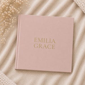 Puede incluir: Un libro de tapa dura cuadrado de color rosa polvoriento con el nombre EMILIA GRACE grabado en letras doradas en la portada. El libro presenta un acabado mate suave y está colocado en el centro, mostrando un diseño limpio y sencillo.