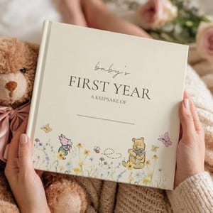 Puede incluir: Un libro de recuerdos para bebés de color crema y forma cuadrada, titulado "baby's FIRST YEAR A KEEPSAKE OF" con ilustraciones de Winnie the Pooh, Piglet, mariposas y flores silvestres en la parte inferior. Junto a un oso de peluche marrón con lazo rosa.