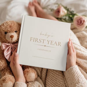 May include: A square white memory book with gold text that reads baby's FIRST YEAR A KEEPSAKE OF with a small heart icon. The book is positioned next to a brown plush teddy bear wearing a light pink satin bow around its neck.