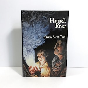 May include: A book cover with a dark background and a blue and white smoke effect. The title "Hatrack River" is in white text and the author "Orson Scott Card" is in white text below. The cover features a young boy holding a glowing orb and an older man with a feathered headdress.