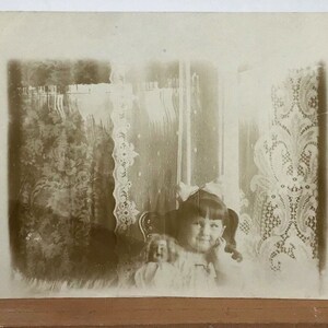 RPPC antico - Ragazza sorridente che tiene una bambola all'interno con tende di pizzo - Cartolina fotografica reale dei primi anni del 1900, foto d'infanzia nostalgica carina