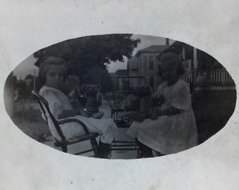 1910 年頃の近所の家のポーチに座っている 2 人のかわいい女の子とテディベアと人形の魅力的な RPPC - アンティークのリアル フォト ポストカード