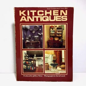May include: A vintage book titled "KITCHEN ANTIQUES" with a maroon cover. The cover features four inset photographs showcasing antique kitchen items. The book's title is in large, bold, white letters. The author and photographer's names are also visible.