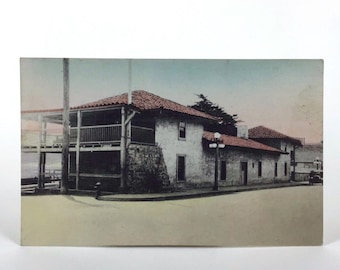 May include: A vintage postcard showing a two-storey building with a red tiled roof and white stucco walls. The building has a large porch with wooden railings and a stone foundation. There is a street in front of the building with a lamppost and a few trees.