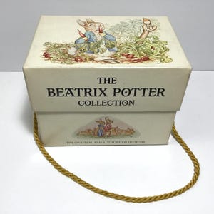 May include: A cream-colored box from "The Beatrix Potter Collection" with a gold rope handle. The box features illustrations of Peter Rabbit and other characters. The text "THE BEATRIX POTTER COLLECTION" is printed on the front.
