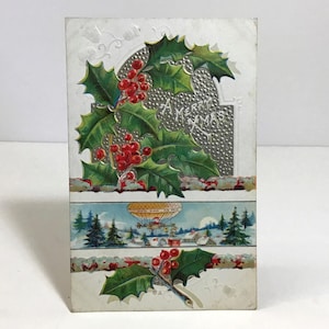 May include: Vintage Christmas postcard with a silver foil background and the text "A Merry Xmas". The postcard features holly leaves and red berries, and a scene of a hot air balloon flying over a snowy winter landscape.