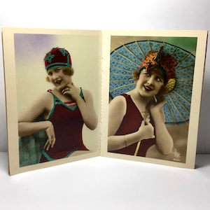 Könnte beinhalten: Vintage Postkarte mit zwei Frauen in Badebekleidung. Eine trägt einen weinroten Badeanzug und eine passende Mütze mit grünen Akzenten. Die andere hält einen blauen Sonnenschirm und trägt ein gemustertes Kopftuch und einen weinroten Badeanzug.