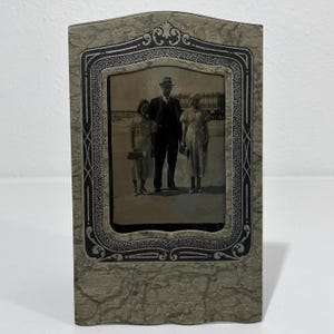 May include: A rectangular picture frame with a textured beige finish and a black decorative border featuring intricate scrollwork. The frame holds a vintage black and white photograph of two individuals standing on either side of a person in a suit.