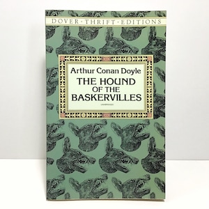 May include: A paperback book titled "The Hound of the Baskervilles" by Arthur Conan Doyle. The cover features a repeating pattern of a snarling dog's head on a green background. The book is part of the Dover Thrift Editions series.