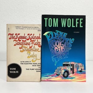 May include: Two books by Tom Wolfe. The left book is titled The Kandy-Kolored Tangerine-Flake Streamline Baby with cream cover and red text. The right book is titled The Electric Kool-Aid Acid Test with a black cover, green text, and a colorful bus.