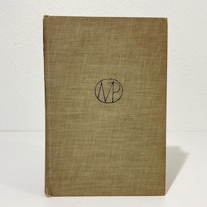 May include: A hardcover book with a tan, textured fabric cover. Centered on the front cover is a black circular emblem containing the stylized letters MP. The book is standing upright, showcasing its rectangular shape and woven material texture.