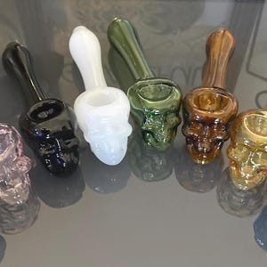 May include: A collection of glass pipes in various colors, including pink, black, white, green, brown, yellow, and blue. Each pipe is shaped like a skull with a bowl and stem.