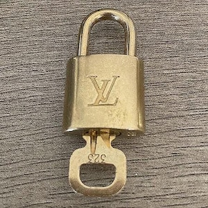 May include: A gold-colored padlock with a key. The padlock has the embossed initials "LV" on the front. The key is attached and has the number "523" engraved on it. The padlock is made of metal.