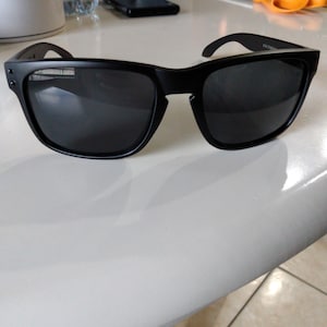 Puede incluir: Gafas de sol negras con lentes oscuras. Las gafas tienen una montura cuadrada y patillas gruesas. Las gafas están sobre una superficie blanca.