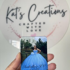 Puede incluir: Un im&aacute;n rectangular con una fotograf&iacute;a de una persona con un vestido de quincea&ntilde;era azul claro. El texto "Isabella's XV 5.17.25" es visible. El fondo incluye un edificio y &aacute;rboles. El im&aacute;n se sostiene frente a un fondo blanco con el texto "Kat's Creations Crafted With Love".