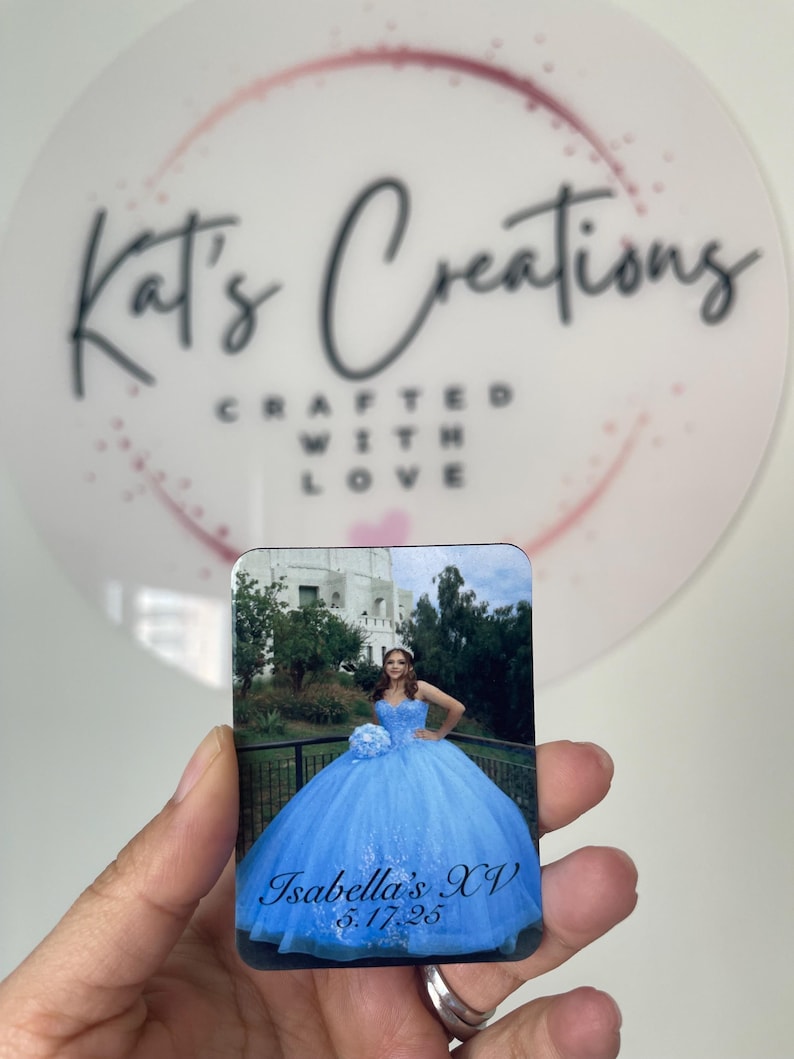 Puede incluir: Un im&aacute;n rectangular con una fotograf&iacute;a de una persona con un vestido de quincea&ntilde;era azul claro. El texto "Isabella's XV 5.17.25" es visible. El fondo incluye un edificio y &aacute;rboles. El im&aacute;n se sostiene frente a un fondo blanco con el texto "Kat's Creations Crafted With Love".
