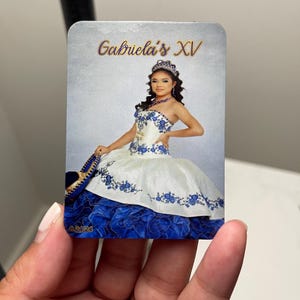 Puede incluir: Tarjeta rectangular con una joven en un vestido de quincea&ntilde;era blanco y azul. El vestido tiene bordados florales y una falda en capas. El texto "Gabriela's XV" est&aacute; escrito en oro. La fecha "8.24.24" es visible.
