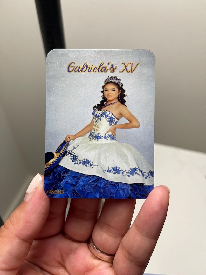 Puede incluir: Tarjeta rectangular con una joven en un vestido de quincea&ntilde;era blanco y azul. El vestido tiene bordados florales y una falda en capas. El texto "Gabriela's XV" est&aacute; escrito en oro. La fecha "8.24.24" es visible.