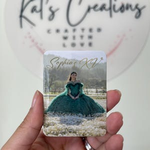 Puede incluir: Un im&aacute;n rectangular con una fotograf&iacute;a de una mujer con un vestido verde de quincea&ntilde;era. El texto "Sophia's XV" est&aacute; escrito en escritura dorada. El fondo incluye un campo de flores blancas. El im&aacute;n est&aacute; sostenido por una mano.