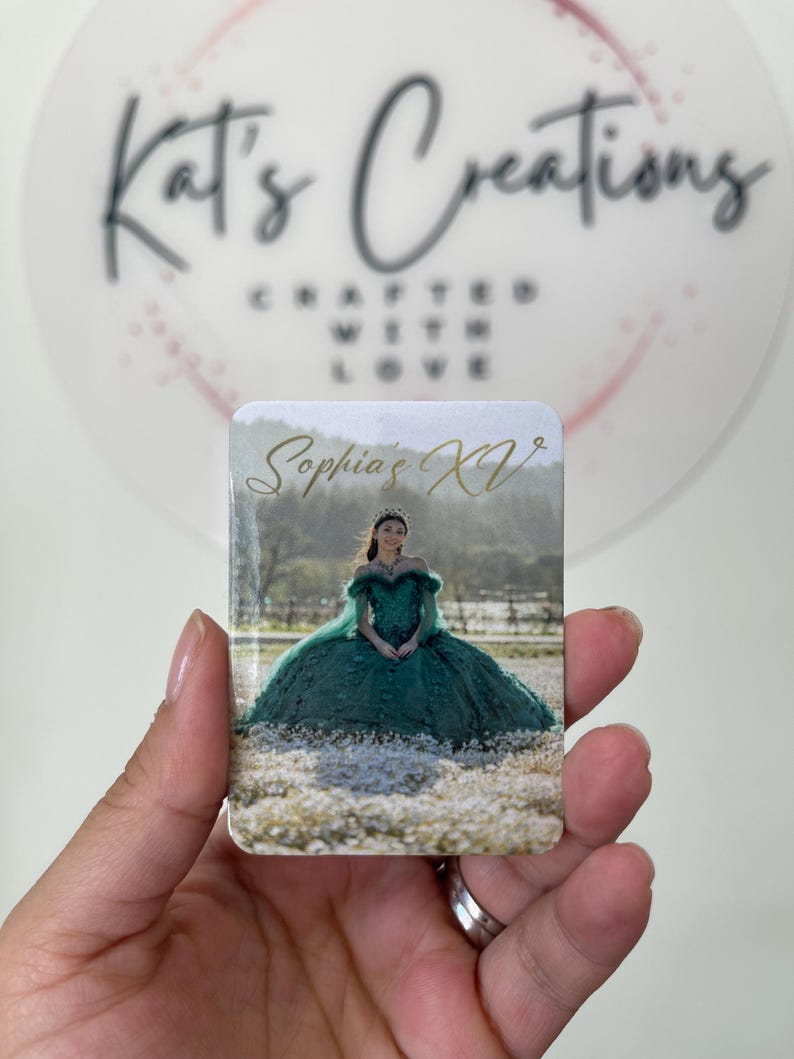 Puede incluir: Un im&aacute;n rectangular con una fotograf&iacute;a de una mujer con un vestido verde de quincea&ntilde;era. El texto "Sophia's XV" est&aacute; escrito en escritura dorada. El fondo incluye un campo de flores blancas. El im&aacute;n est&aacute; sostenido por una mano.