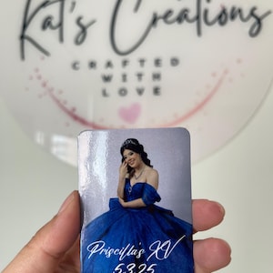 Puede incluir: Un im&aacute;n rectangular con una joven con un vestido de baile azul real. El im&aacute;n tiene el texto "Priscilla's XV 5.3.25" en escritura blanca. El fondo es una imagen borrosa de un c&iacute;rculo blanco con el texto "Kat's Creations Crafted With Love."