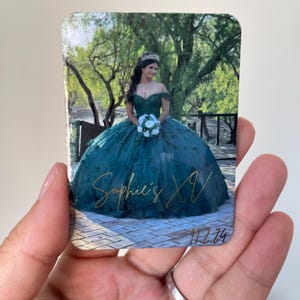 Puede incluir: Un im&aacute;n rectangular con una joven en un vestido de quincea&ntilde;era verde azulado, sosteniendo un ramo. El im&aacute;n tiene el texto "Sophie's XV" en escritura dorada, con la fecha "11.2.24" debajo. El fondo muestra un puente y &aacute;rboles.