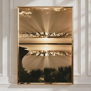 Peut inclure: Une impression d'art mural encadrée représentant un paysage sépia d'un lac calme au coucher du soleil. Le soleil se trouve bas à l'horizon derrière une bande horizontale de nuages, projetant des rayons lumineux vers le haut sur l'eau.