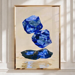 May include: A framed wall art print featuring two stacked, translucent, deep blue crystalline geometric shapes floating above a third, flatter blue crystal form that rests on a light beige, rippled, water-like surface with soft reflections.