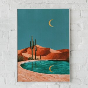 May include: A rectangular wall art print featuring a desert landscape with orange sand dunes, two tall green cacti, and a turquoise pool in the foreground. A golden crescent moon is visible in the teal sky and reflected in the water below.