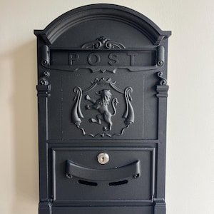 Puede incluir: Buzón de metal negro con un escudo de león y la palabra "POST" en la parte delantera. El buzón tiene una ranura para el correo y una pequeña puerta con una cerradura.