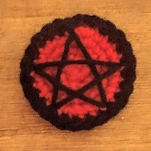 Puede incluir: Un pentagrama rojo tejido a crochet con un contorno negro. El pentagrama está rodeado por un círculo negro tejido a crochet.