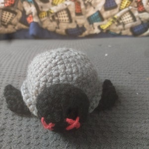 Op de afbeelding: Een grijze gehaakte pinguïn met zwarte ogen en een rode snavel. De pinguïn ligt op zijn buik met zijn vleugels uitgespreid.