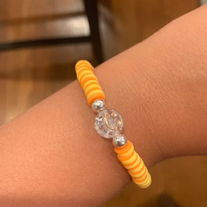 May include: A yellow and orange striped bracelet with a silver charm. The charm is a clear circle with a silver design inside.