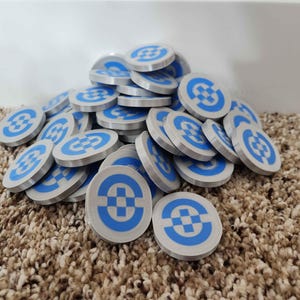 May include: A pile of round, silver-colored tokens with a blue and white geometric design. The tokens are stacked on a brown carpet, with a white wall in the background. The tokens appear to be made of plastic or metal.