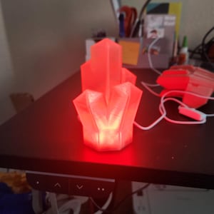 May include: A red, geometric crystal-shaped lamp is illuminated on a dark surface. The lamp is connected to a white cord. The lamp's design features multiple facets, creating a unique, glowing effect. The background includes a desk and other office items.