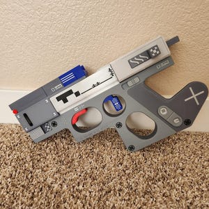 May include: A gray and white toy gun with a red trigger, blue accents, and a plus sign on the handle. The gun has a retro video game aesthetic with various buttons and pixelated designs.