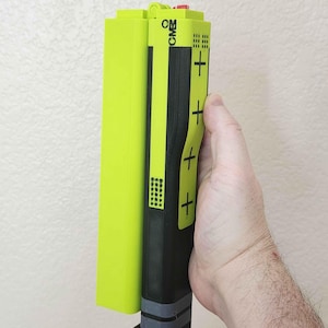 May include: A 3D-printed device with a neon green rectangular component attached to a black, rectangular body. The body has yellow accents, plus signs, and the letters "CMB". A red lever is visible at the bottom. The device is held by a hand.