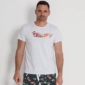 May include: A white t-shirt with a Christmas-themed graphic of Santa Claus in a sleigh pulled by reindeer. The person is wearing matching green and white Christmas-themed pajama shorts.