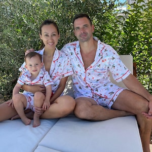 May include: A family of three wearing matching white pajamas with a red trim and a festive print. The pajamas feature a variety of holiday-themed illustrations, including Santa Claus, reindeer, and snowmen. The pajamas are personalized with the name "Amy" on the woman's shirt.