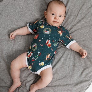 May include: A baby wearing a green and white Christmas-themed onesie with a white trim. The onesie has a pattern of presents, candy canes, and reindeer. The baby is lying on a gray round mat.