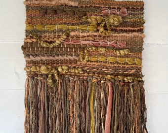May include: A woven wall hanging with a wooden dowel and off-white cords for hanging. The textile features a mix of textures and colors, including shades of brown, green, and pink. The bottom edge has long, flowing fringe in similar tones.