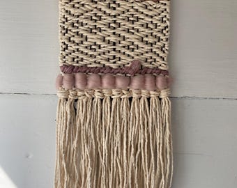 May include: A woven wall hanging with a geometric pattern in beige and pink. The hanging has a fringe of beige yarn and a wooden dowel at the top.